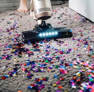 A vacuum cleaner is being used to clean colorful confetti off a grey carpet. The vacuum is silver with a lit nozzle showing the confetti being sucked in. A pair of bare feet is visible behind the device, indicating a person is operating it.