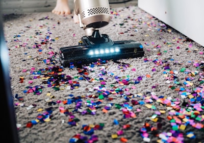 A vacuum cleaner is being used to clean colorful confetti off a grey carpet. The vacuum is silver with a lit nozzle showing the confetti being sucked in. A pair of bare feet is visible behind the device, indicating a person is operating it.