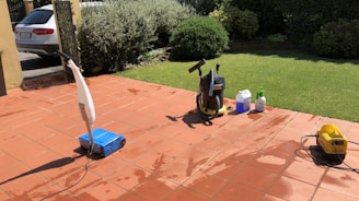 A sunlit patio with terracotta tiles is adorned with cleaning equipment, including a wet and dry vacuum, containers of cleaning solutions, and a pressure washer. A portion of a white car is visible near a hedge-lined driveway, while lush green vegetation and a manicured lawn provide a backdrop.