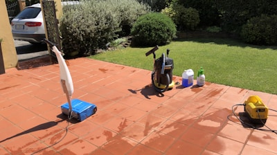 A sunlit patio with terracotta tiles is adorned with cleaning equipment, including a wet and dry vacuum, containers of cleaning solutions, and a pressure washer. A portion of a white car is visible near a hedge-lined driveway, while lush green vegetation and a manicured lawn provide a backdrop.