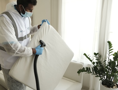 A person wearing protective clothing, a face mask, and gloves is cleaning a white cushioned furniture piece using a vacuum cleaner. The setting appears to be indoors with bright natural light streaming in through large windows. A green potted plant is placed next to a white sofa, adding a touch of nature to the clean and tidy environment.