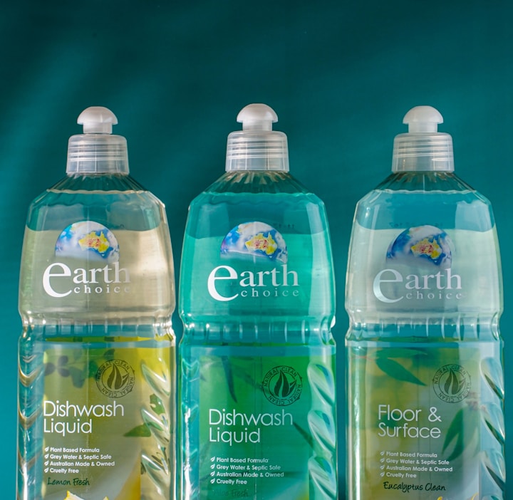 Three bottles of Earth Choice cleaning products are lined up on a wooden surface. The bottles have liquid inside and feature colorful labels with natural imagery like lemons and flowers. Each bottle is topped with a white cap, and the background is a solid teal color.