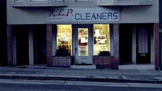 A dry cleaning shop named 'HLP Cleaners' is located on a dimly lit street. The store has a prominent sign above the entrance, and its windows are illuminated with a warm yellow light. A sign on the door lists services such as dry cleaning, laundry, and suede & leather cleaning. The building exterior is simple with a combination of dark tiles and light-colored upper walls. The entrance is flanked by two closed doors, likely of neighboring establishments.