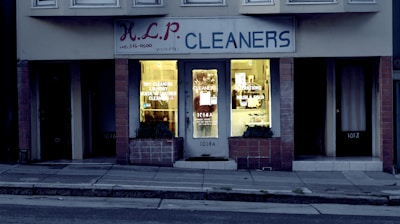 A dry cleaning shop named 'HLP Cleaners' is located on a dimly lit street. The store has a prominent sign above the entrance, and its windows are illuminated with a warm yellow light. A sign on the door lists services such as dry cleaning, laundry, and suede & leather cleaning. The building exterior is simple with a combination of dark tiles and light-colored upper walls. The entrance is flanked by two closed doors, likely of neighboring establishments.