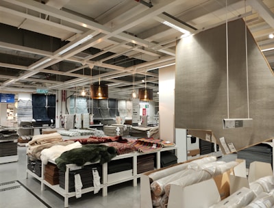 A retail store interior featuring a large selection of rolled-up carpets and rugs displayed on shelves and tables. Several hanging lamps provide lighting to the area, and a variety of carpet styles and colors are visible. The shelving units are neatly organized with packaged flooring products, and a large sign displaying product information is visible.