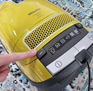 A yellow vacuum cleaner with a control panel featuring various icons for settings. A finger is pressing a button on the panel. The vacuum is placed on a patterned carpet with shades of blue, white, and black.