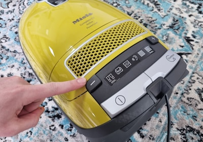 A yellow vacuum cleaner with a control panel featuring various icons for settings. A finger is pressing a button on the panel. The vacuum is placed on a patterned carpet with shades of blue, white, and black.