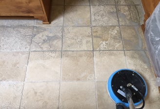 A tiled floor with visible dirt and cleaning solution being cleaned by a circular floor cleaning machine. The tiles are light brown with noticeable dirt in the grout lines. The cleaning machine has a blue and black circular head connected to a metal handle and hose.