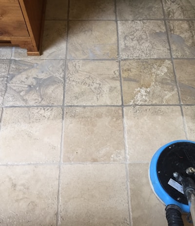 A tiled floor with visible dirt and cleaning solution being cleaned by a circular floor cleaning machine. The tiles are light brown with noticeable dirt in the grout lines. The cleaning machine has a blue and black circular head connected to a metal handle and hose.