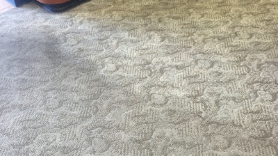 A patterned carpet covers most of the floor, with a yellow floor cleaning machine partially visible on the right. The pattern on the carpet appears to be a mix of geometric and abstract shapes. A section of a wooden counter with a curved design is on the left side.