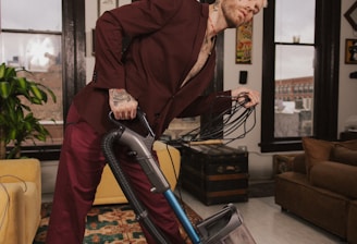 A person wearing a maroon suit is leaning back while vacuuming a rug in a neatly arranged room. The room features large windows with black frames, a plant, and various pieces of furniture such as a yellow armchair and a brown sofa. The walls are adorned with framed pictures and artwork.