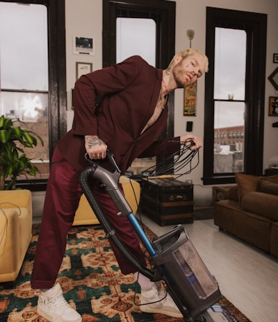 A person wearing a maroon suit is leaning back while vacuuming a rug in a neatly arranged room. The room features large windows with black frames, a plant, and various pieces of furniture such as a yellow armchair and a brown sofa. The walls are adorned with framed pictures and artwork.